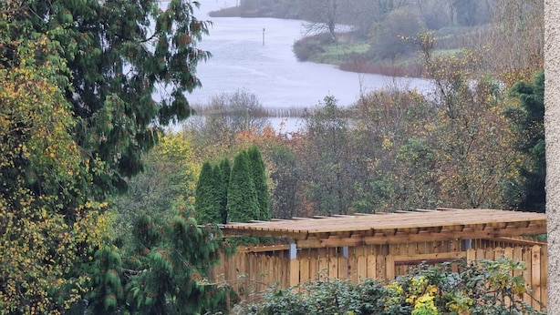 A wooden cabin with a sloped roof, surrounded by dense foliage and a river in the background. - 3-beds, Sleep 7 With Garden & Private Parking (Carrick-on-Shannon)