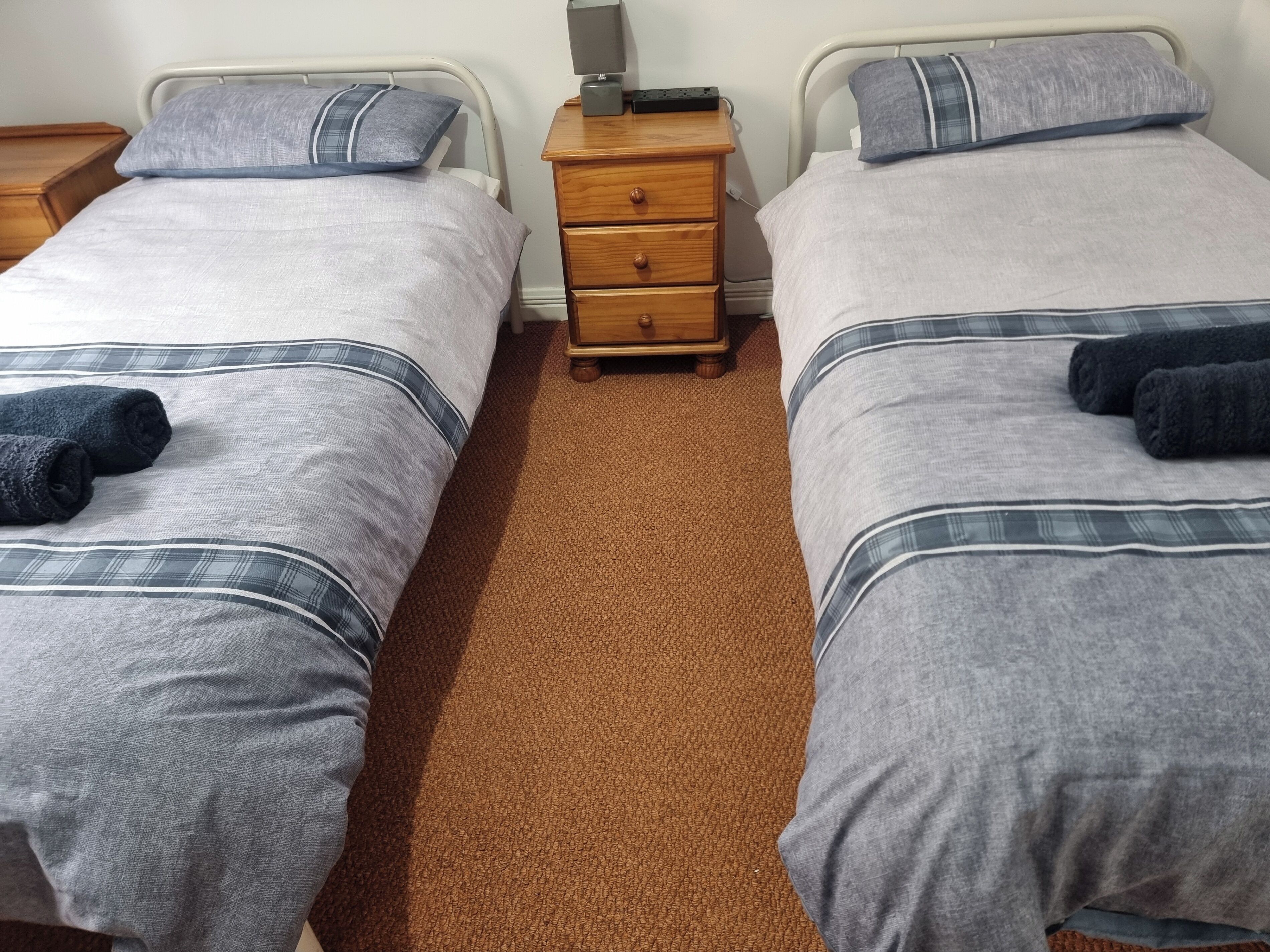 Habitación doble con dos camas, cada una con una manta gris y una almohada azul. Hay una mesita de noche de madera entre las camas con una lámpara y un enchufe.