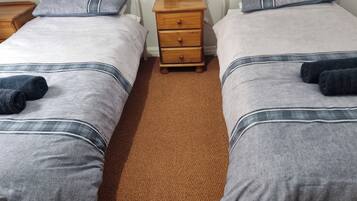 Habitación doble con dos camas, cada una con una manta gris y una almohada azul. Hay una mesita de noche de madera entre las camas con una lámpara y un enchufe.