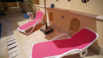 Deux chaises longues blanches avec des coussins roses, une petite table et une plante en pot, le tout placé dans un espace extérieur ensoleillé doté d’un sol carrelé et d’un mur jaune agrémenté de fenêtres décoratives.