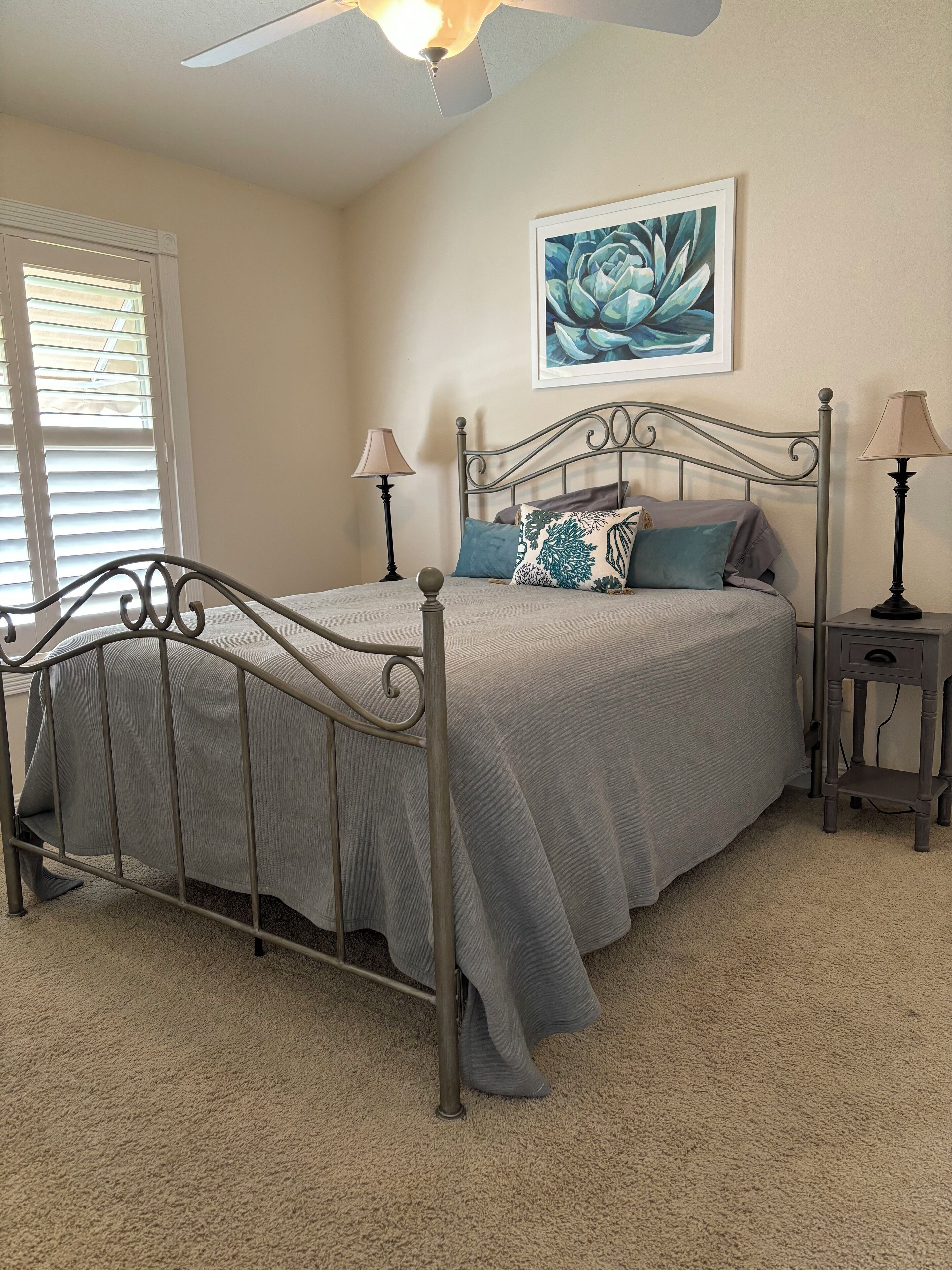 A bedroom with a metal bed frame, a gray bedspread, two bedside lamps, a ceiling fan, and a wall-mounted artwork.