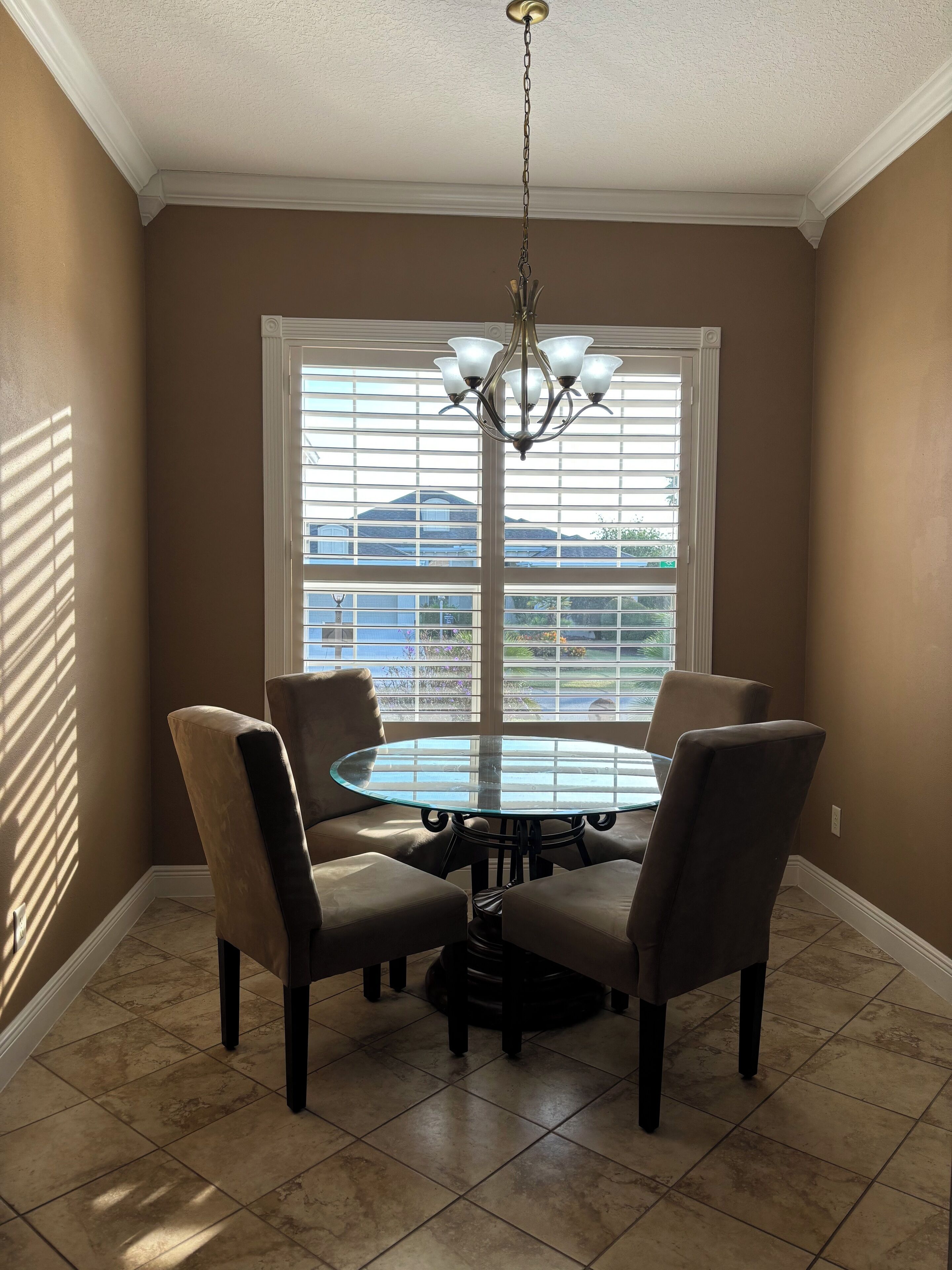 A dining area with a round glass table, four chairs, a chandelier, and a large window with blinds.