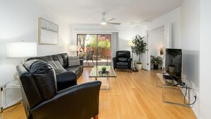 A modern living room with a leather sofa, a glass coffee table, a black armchair, and a TV on a stand. There is a ceiling fan, a door leading to a balcony, and several potted plants. - CASA FAMILIA - 1st floor 2-bedroom unit (Phoenix)