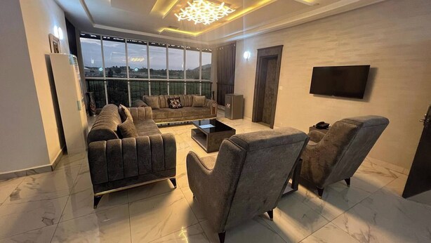 A modern living room with a sectional sofa, armchairs, a coffee table, and a TV mounted on the wall. - Jay Hiltin Hotel (Kaduna)