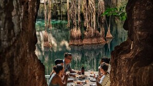 Un groupe de personnes est réuni autour d’une table dans un endroit qui ressemble à une grotte, avec des racines pendantes et une vue sur une piscine naturelle.
