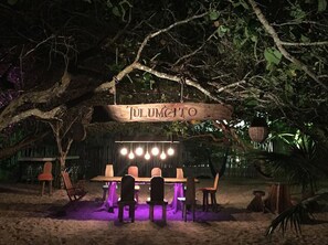 An outdoor dining area with a wooden table and chairs under a tree, illuminated by hanging lights. - Beachfront Villa - Padel & Glamping at LAHOS (Chicxulub)