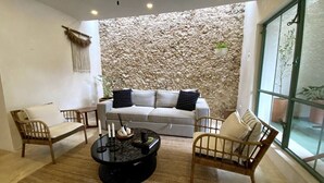 A cozy living room with a stone wall, a white sofa, two wooden chairs, and a coffee table with candles. - Casa Verde Maya Downtown by LAHOS (Mérida)