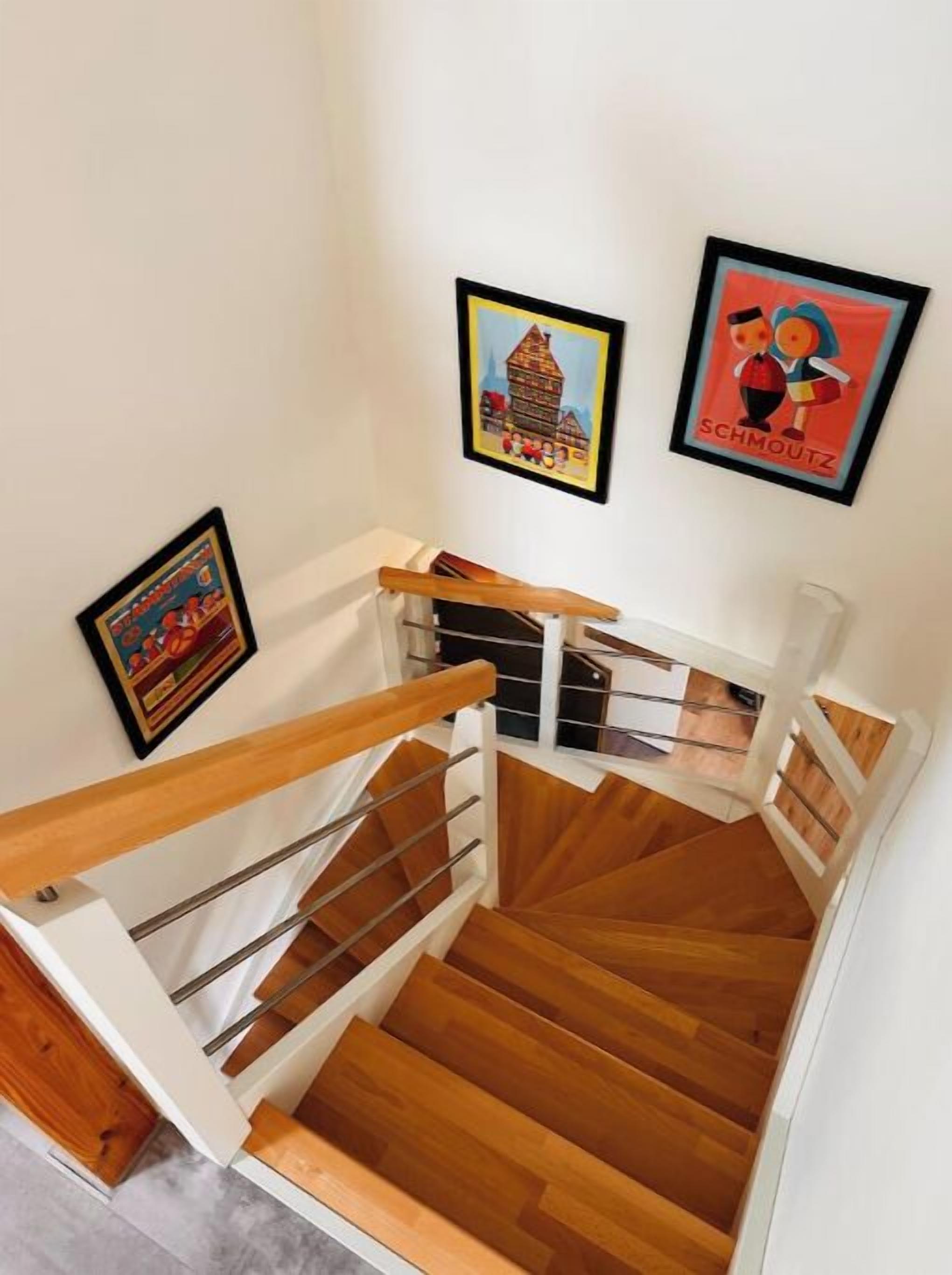 Un escalier en bois avec des rampes en métal et des murs blancs. Trois tableaux encadrés sont accrochés au mur.