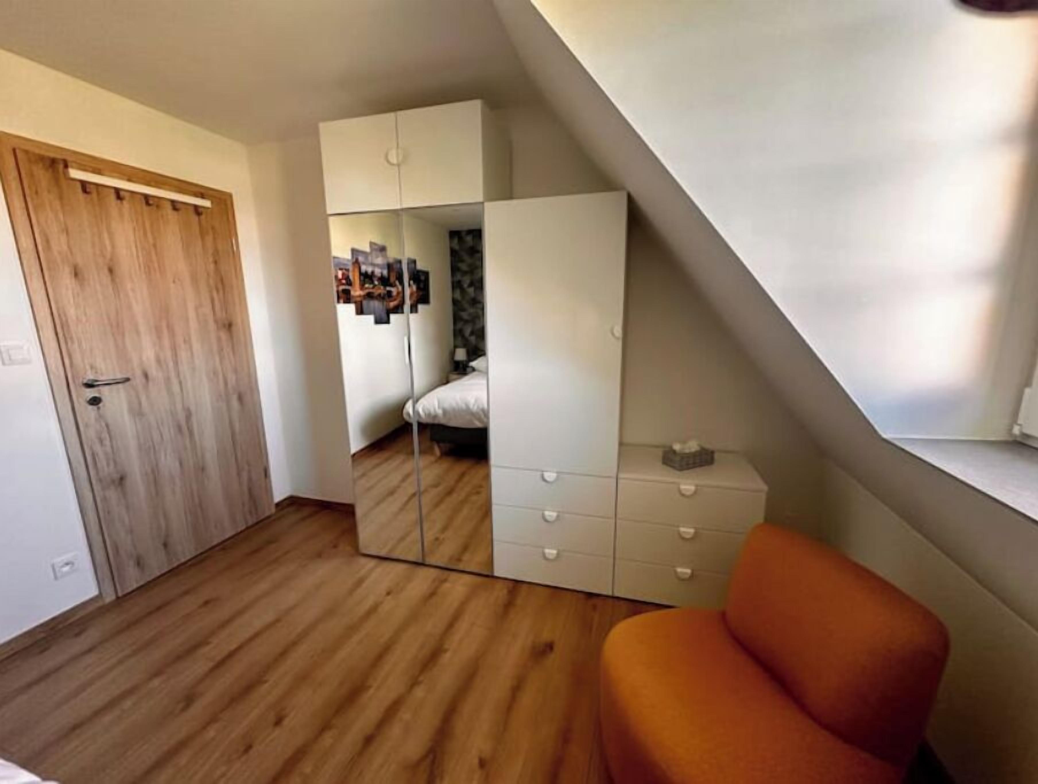 A bedroom with a wooden door, a bed, a wardrobe, and a small orange chair.