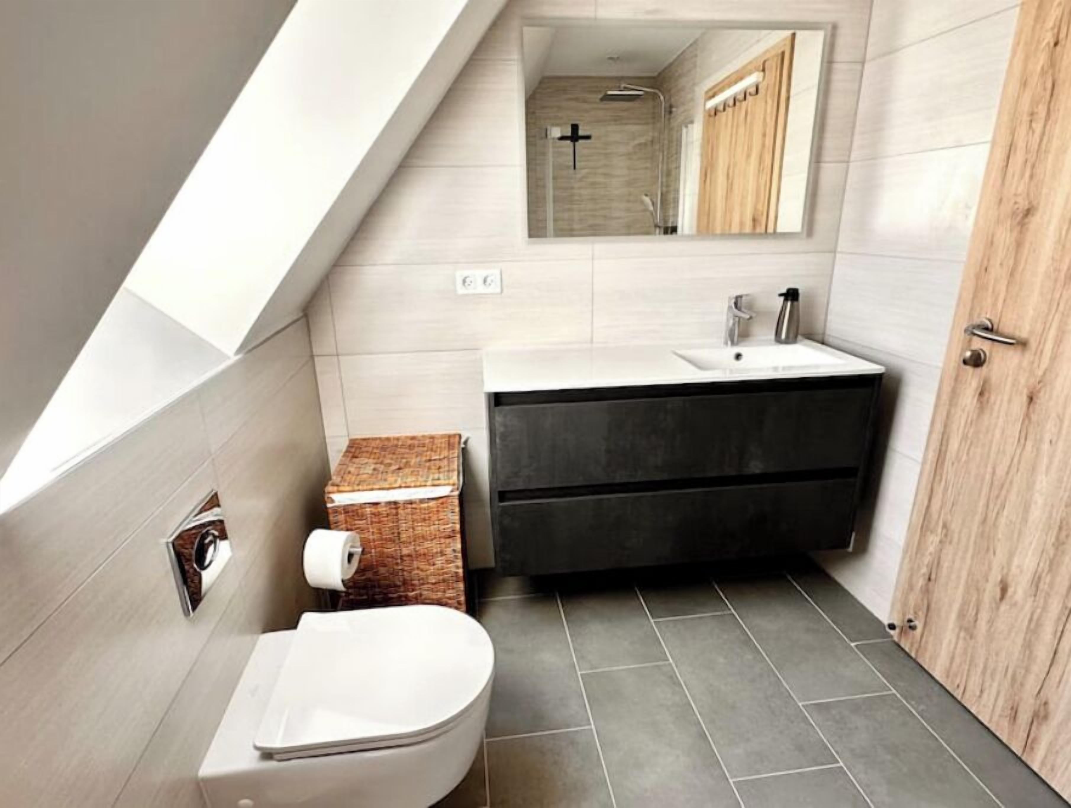 Een moderne badkamer met een schuine balkenlaag, toilet, wastafel en spiegel.
