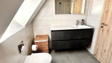 Een moderne badkamer met een schuine balkenlaag, toilet, wastafel en spiegel.
