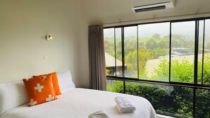 A hotel room with a large bed, an air conditioning unit, a window with a view of greenery and buildings, and a wall-mounted light. - Delatitie Apartments -Cedar 1 Bedroom Apartment - Merrijig (Merrijig)
