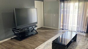 Una sala de estar con un televisor de pantalla plana sobre un soporte negro, una mesa de centro de vidrio y una alfombra estampada.