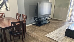 Una sala de estar con una mesa de comedor de madera, cuatro sillas de madera, un televisor de pantalla plana sobre un mueble bajo y una alfombra estampada.