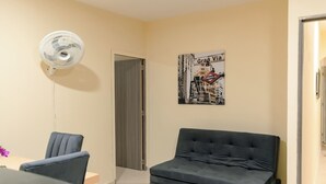 Une pièce avec un canapé sombre, un ventilateur blanc, une table à manger avec des chaises et une affiche accrochée au mur.
