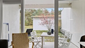Une pièce dotée d’une porte-fenêtre coulissante qui donne sur une terrasse extérieure. À l’intérieur, on trouve une table blanche, une cafetière noire et un vase rempli de fleurs rouges. Une chaise en osier est placée devant la table.