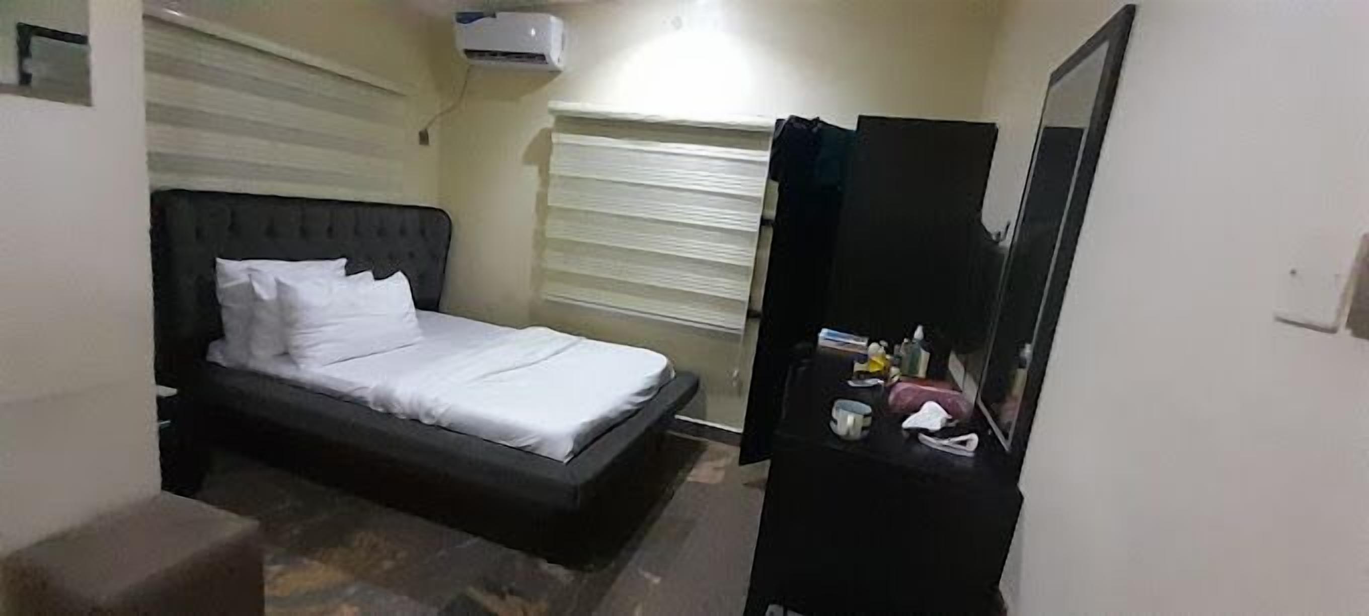 Quarto com cama, mesa de cabeceira com espelho e janela com persianas.