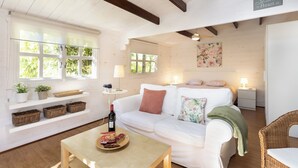 Un salón acogedor con un sofá blanco, una mesa de centro de madera y una silla de mimbre. Hay estanterías con plantas y cestas, además de un cuadro en la pared.