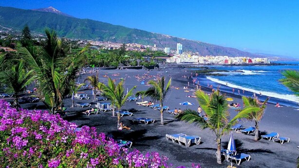 Una playa con tumbonas y palmeras, un pueblo costero al fondo y una montaña.
