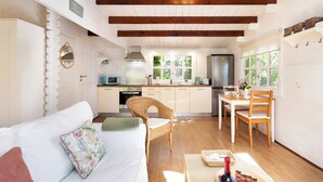 Una habitación acogedora con un sofá blanco, una silla de mimbre, una mesa de comedor con sillas, una zona de cocina con refrigerador y una ventana con vistas a la vegetación.