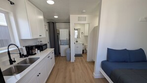 A compact kitchen with white cabinets, a microwave, and a coffee maker. There is a small laundry area with a washing machine and a sink. The room has a wooden floor and a bed with dark blue bedding. - The CB&M Myton Minis  (Myton)