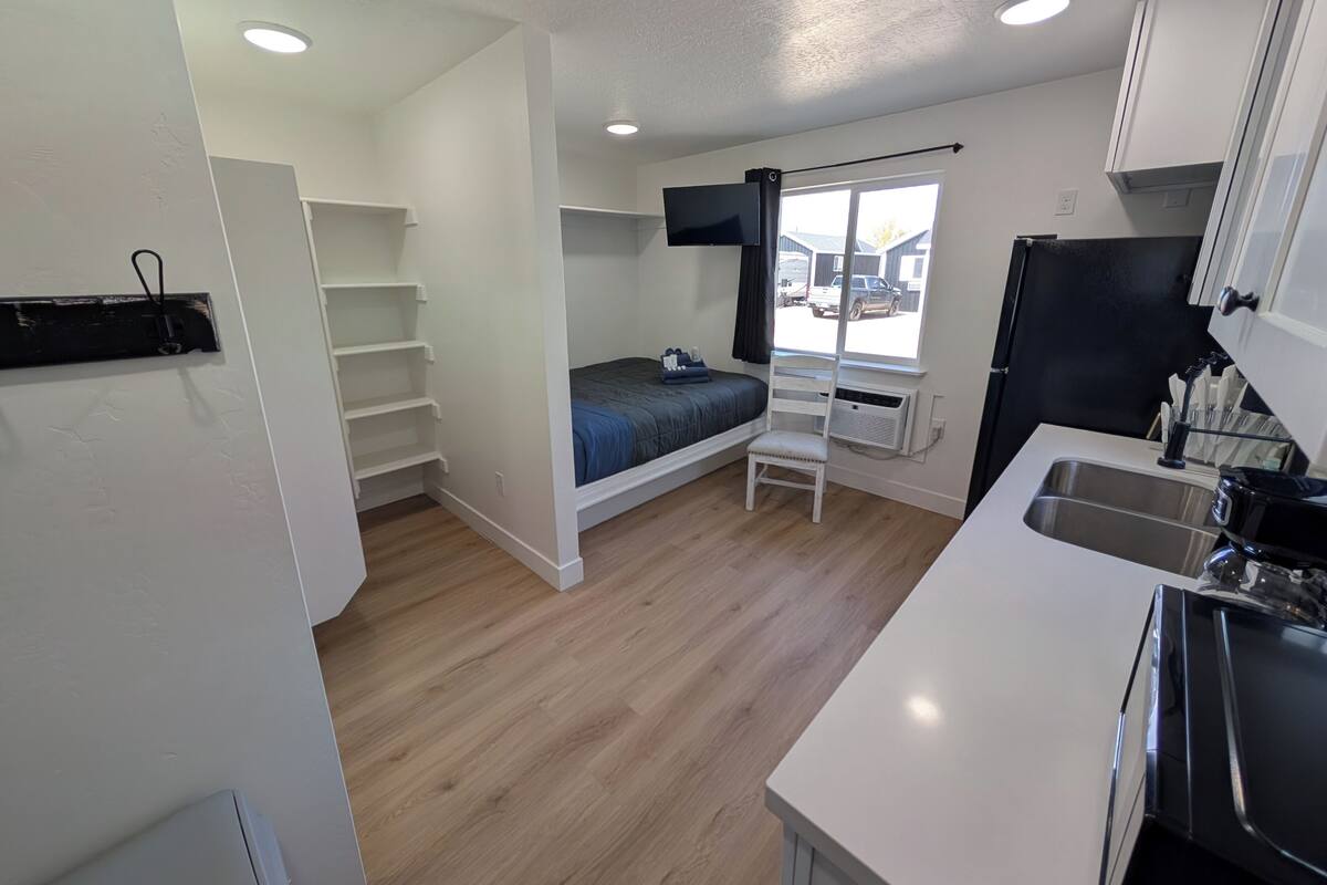 A compact room with a bed, a small desk, a chair, a window, and a kitchenette.