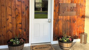 A wooden door with a white frame, a welcome mat, two potted plants, and a sign for Rustic Roots Rentals. - Route 66 Rails to Trails home with Central AC and WIFI (Shippenville)
