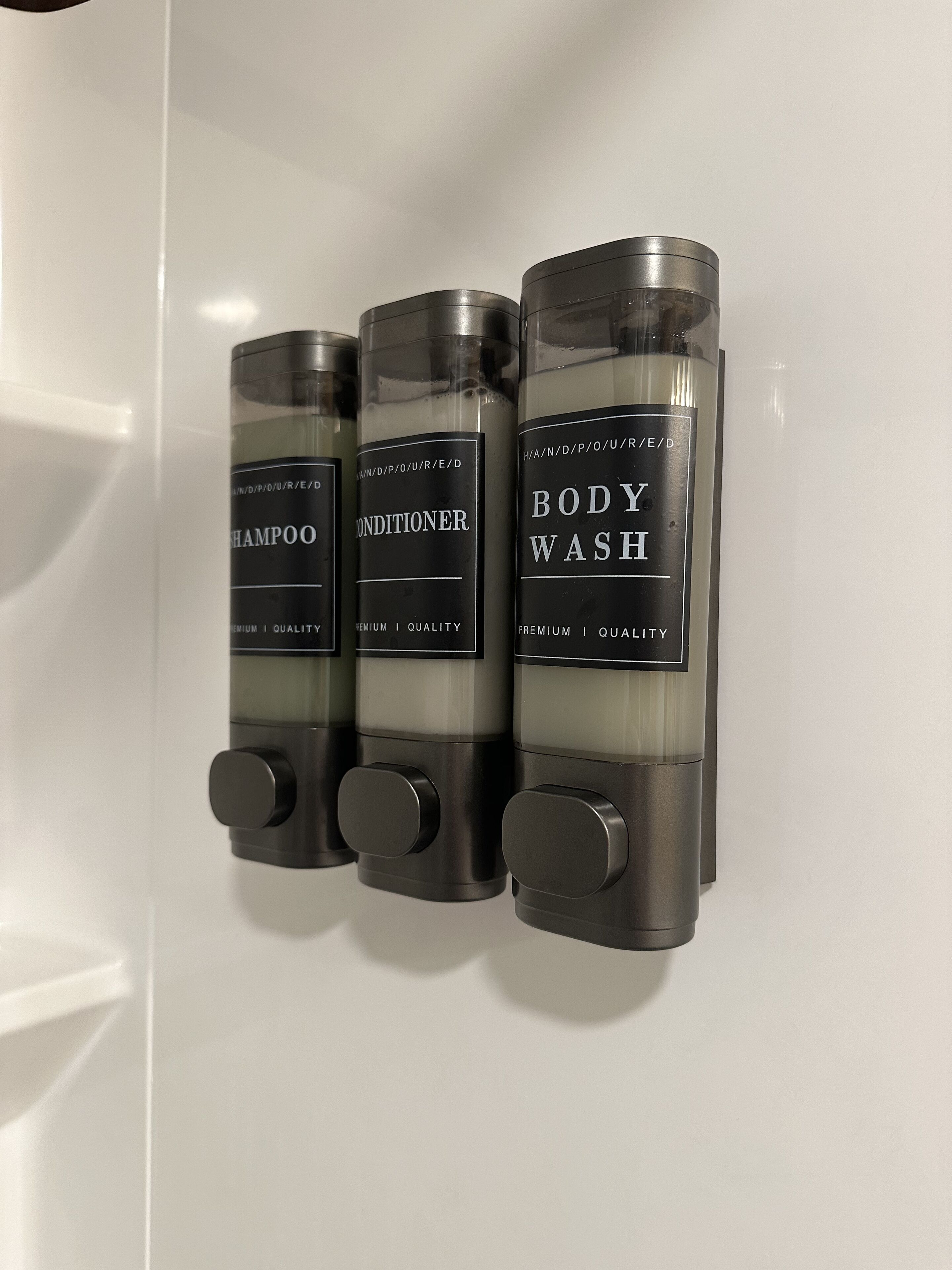 Trois flacons de shampoing, d’après-shampoing et de gel douche, fixés sur un mur blanc.