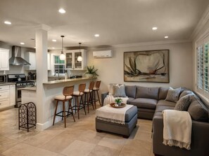 A modern kitchen with white cabinets, a central island with bar stools, a dining area with a gray sofa and patterned pillows, and a large abstract painting on the wall. - Fire Pit & Game Room 4 Beds Near Beach, LAX & SoFi (Lawndale)