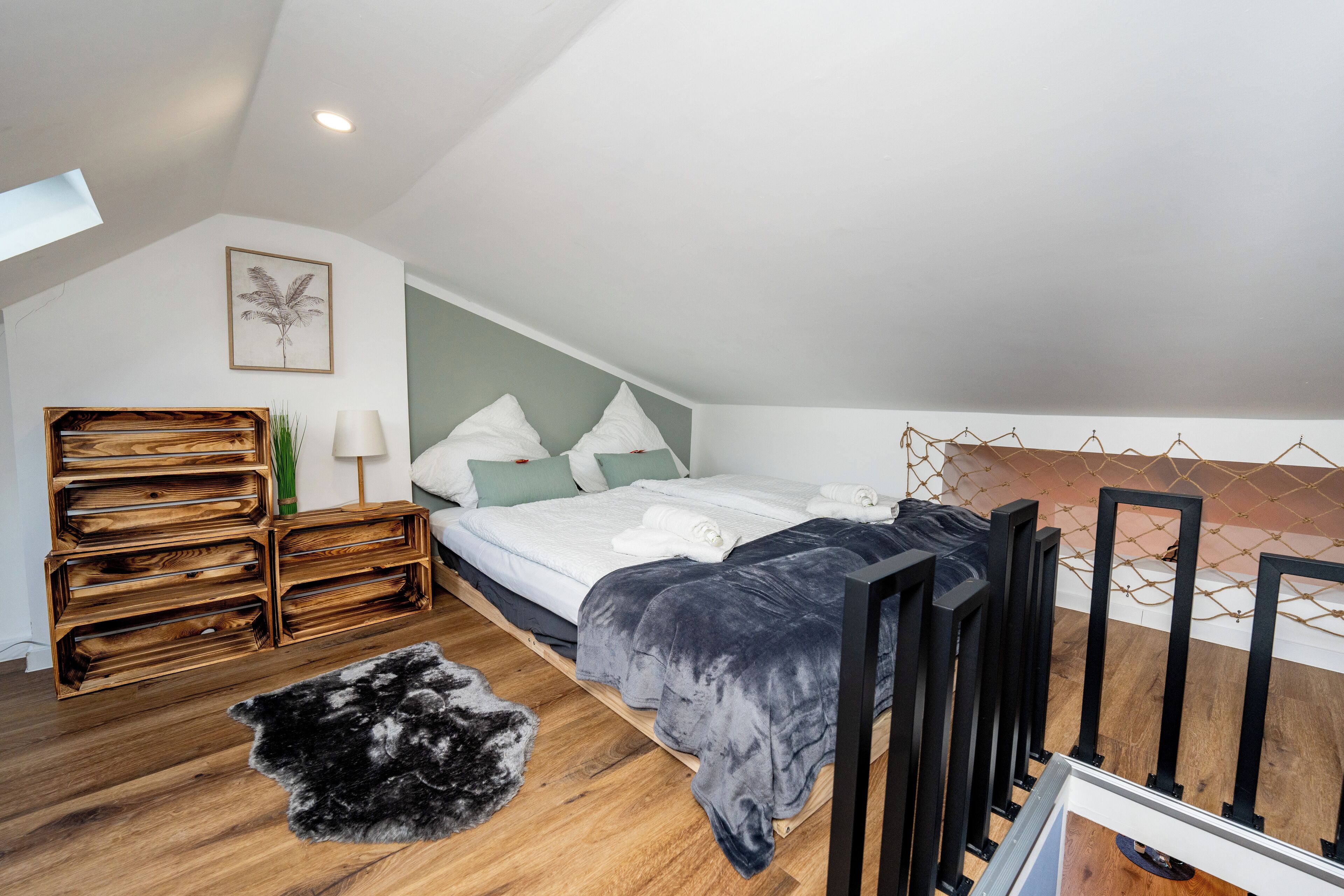 A bedroom with a sloped ceiling, a bed with a grey duvet, wooden crates, a black and white rug, and a framed plant on the wall.