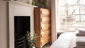 Une chambre avec une cheminée, une commode en bois, un lit recouvert d’une couette blanche et une fenêtre donnant sur des immeubles.