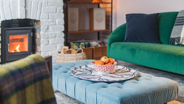 Un salón acogedor con chimenea, un sofá verde y un otomano azul con un frutero.