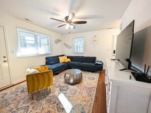 Un salon comprenant un canapé d’angle, un fauteuil jaune, une table basse ronde, un ventilateur de plafond et un téléviseur à écran plat posé sur un meuble blanc.