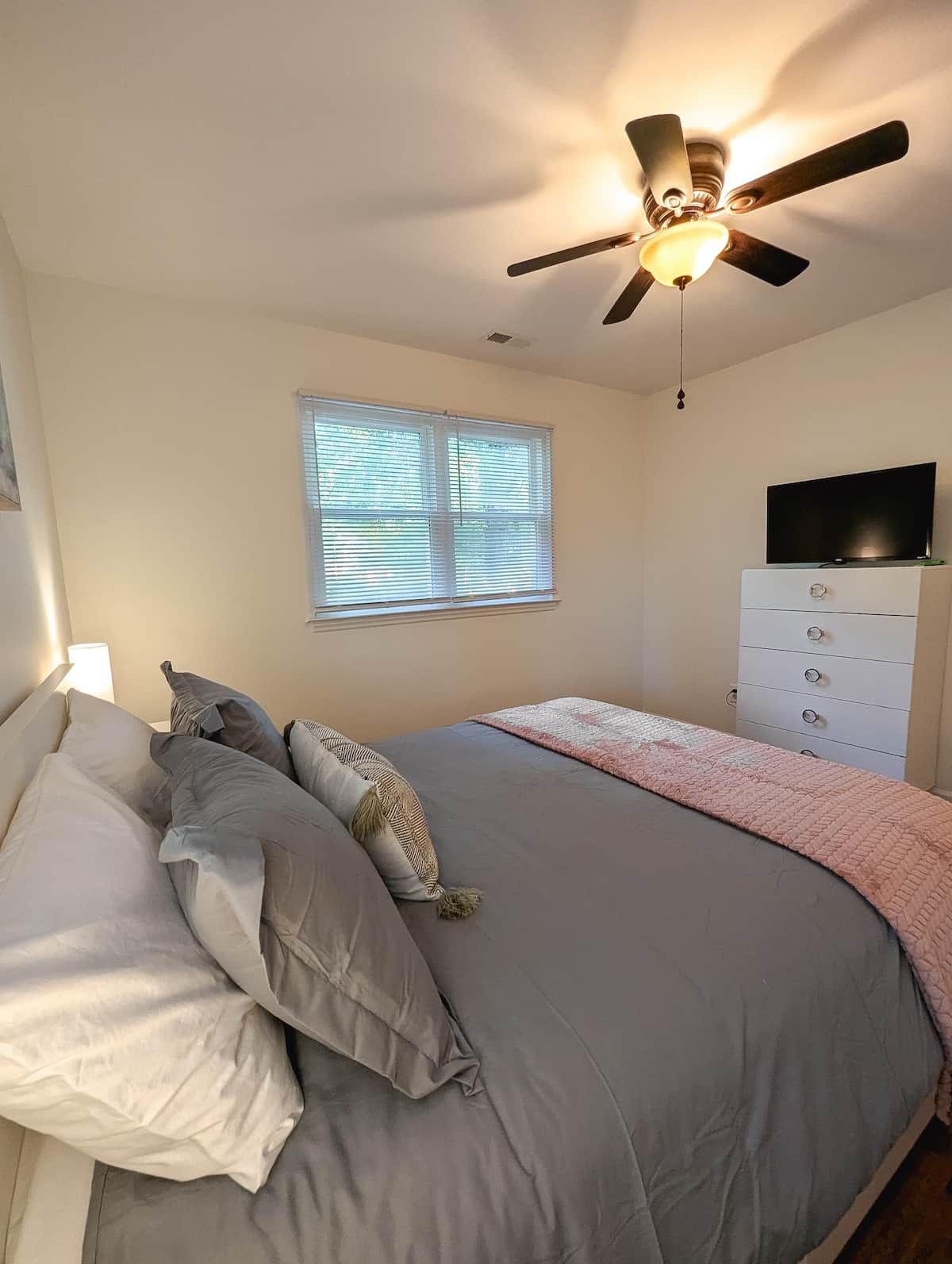 Une chambre à coucher avec un lit, un ventilateur de plafond, une fenêtre avec des stores, un téléviseur posé sur une commode et une lampe de chevet.