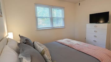 Une chambre à coucher avec un lit, un ventilateur de plafond, une fenêtre avec des stores, un téléviseur posé sur une commode et une lampe de chevet.