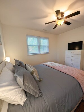 A bedroom with a bed, a ceiling fan, a window with blinds, a TV on a dresser, and a bedside lamp. - Stylish One-Level Near RDU | Cozy & Modern (Morrisville)