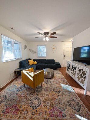 Ein Wohnzimmer mit einem dunkelgrauen Sofa, einem gelben Sessel, einem runden Couchtisch, einem gemusterten Teppich, einem Deckenventilator, einem Fernseher auf einem weißen Schrank und einer Tür im Hintergrund.