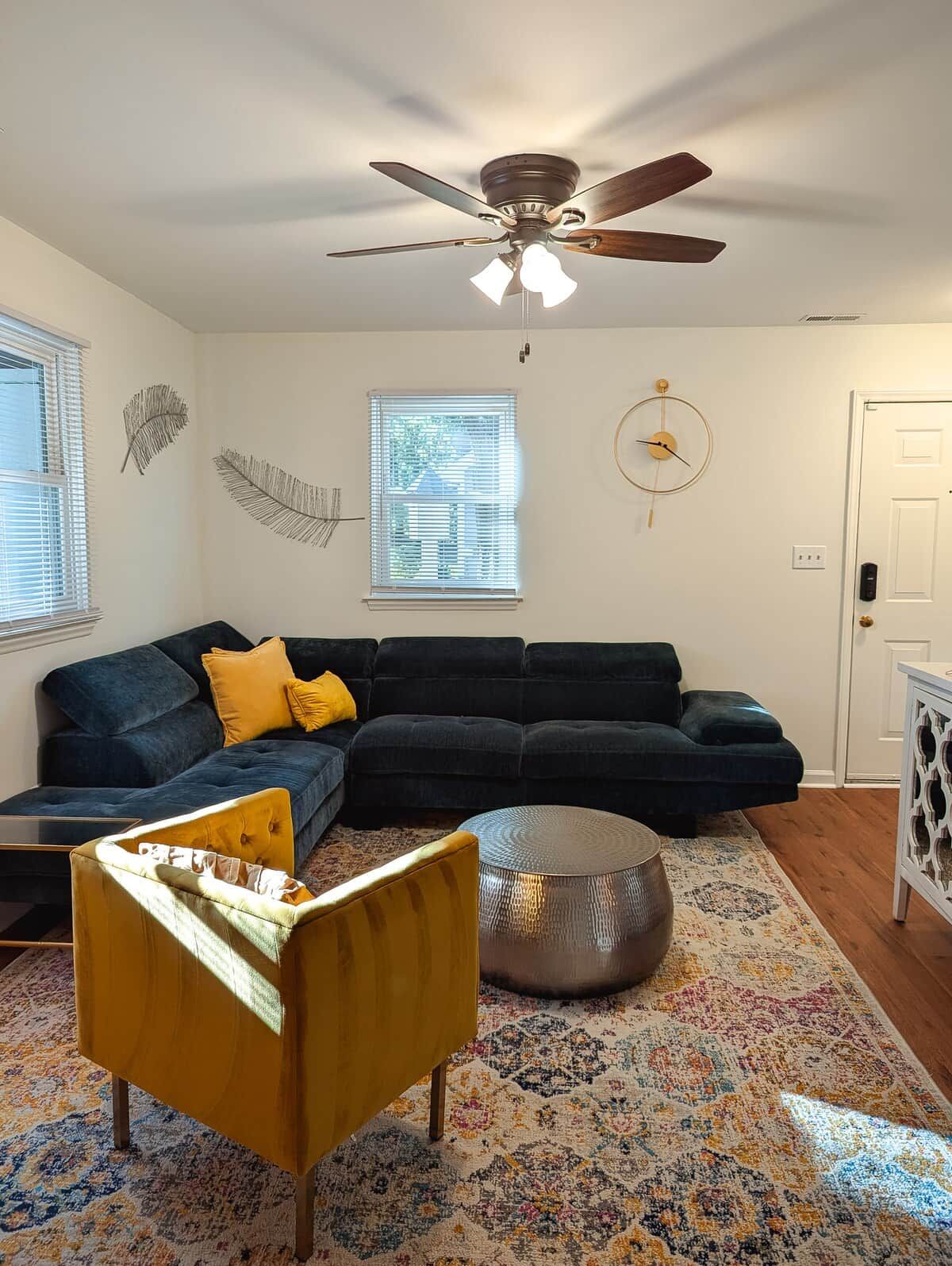 A living room with a dark blue sectional sofa, a yellow armchair, a round ottoman, a ceiling fan, and a patterned rug.