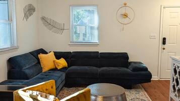 A living room with a dark blue sectional sofa, a yellow armchair, a round ottoman, a ceiling fan, and a patterned rug.