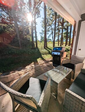 Eine Terrassengruppe im Freien mit Korbmöbeln, einem Grill und Blick auf Bäume und ein Gebäude im Hintergrund.