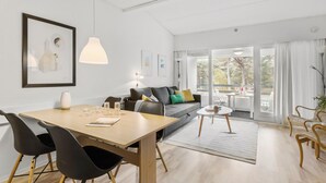 A modern living room with a wooden dining table, black chairs, a grey sofa with colorful cushions, and a large window offering a view of trees. - 1 bedroom lovely apartment in Nexø (Nexø)