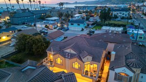 A residential area with houses featuring red-tiled roofs, a prominent house with a balcony, and a view of the ocean and hills in the background. - Two Homes-One block to Beach! (Pismo Beach)