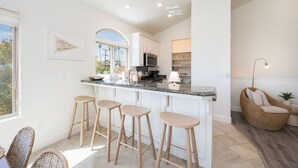 A modern kitchen with a central island, bar stools, and a wicker chair. - Two Homes-One block to Beach! (Pismo Beach)