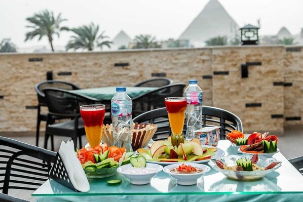 A table set with various dishes, drinks, and a salad, with a stone wall and palm trees in the background. - Night Pyramids Inn (Giza)