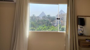 Ein Hotelzimmer mit Blick auf die Pyramiden, ein Fenster mit Vorhängen, eine Klimaanlage, ein Fernseher auf einem Holzboden und ein Nachttisch mit einer Fernbedienung.