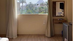 Ein Hotelzimmer mit großem Fenster und Blick auf die Pyramiden, Holzboden, Bett, Minibar und Spiegel.