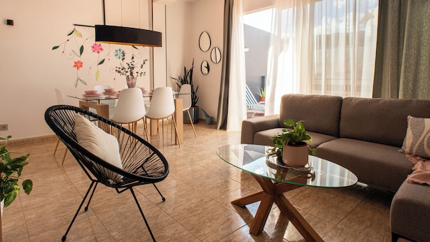 A modern living room with a glass coffee table, a sofa, a dining area with a table and chairs, and a large window with curtains.