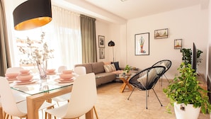 A modern living room with a glass dining table, white chairs, a sofa, a black chair, and a potted plant.