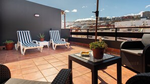 A terrace with two striped lounge chairs, a small table, potted plants, and a view of a cityscape.