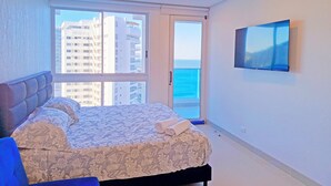 A bedroom with a bed, a TV mounted on the wall, and a view of the sea through a large window. - Exclusive Ocean View Apartment - Reserva del Mar (Santa Marta)